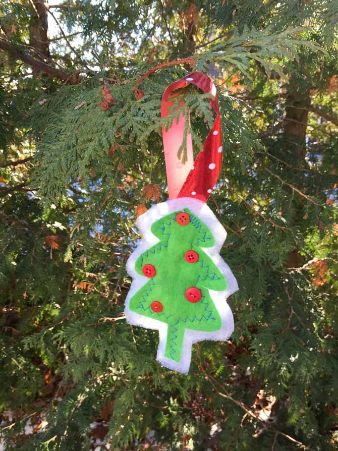 Small tree ornament made from green flannel with small red buttons on it hanging from a cedar tree.
