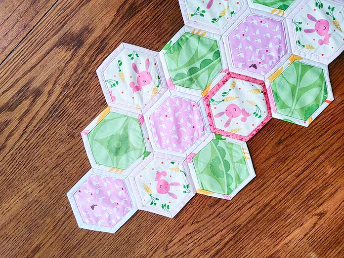 Hexie Table Runner showing green floral, small white bunnies on pink fabric and larger pink bunnies surrounded by orange carrots on small inside hexies. The hexies are framed with white and pink borders.  