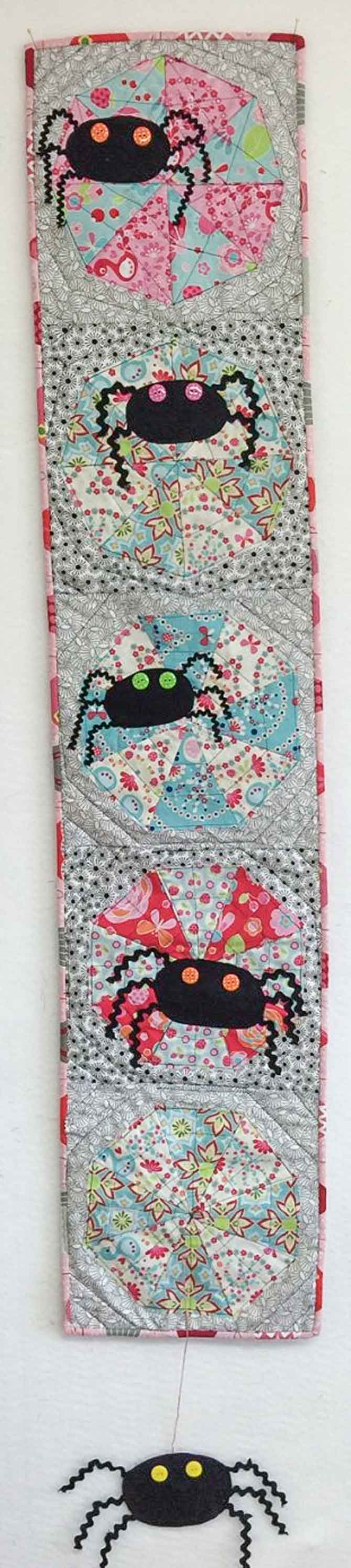 Spider wall hanging with five vertical blocks each having a cobweb in them and the top four having a spider in them and the last block having a spider hang off the quilt hanging by a thread.