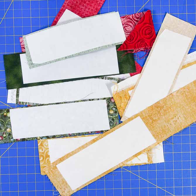 white paper which is fusible web on back of assorted green, red & gold pieces of fabric on a blue cutting mat