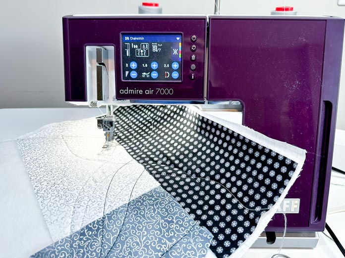 A white and burgundy serger with an extension table supporting a quilt sandwich made of white, gray, and black fabrics, and batting used in the runner to test the thread colours, and thread tension, in chain stitch quilting.