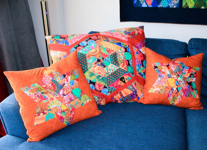 Two smaller 6-point star pillows on either side of a matching oversize diamond hexagon pillow in orange and matching colorful fabrics laid out on a blue couch