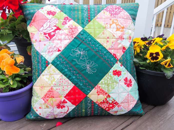 Quilted cushion in red and green colors using 4 nine-patch squares and green stitched setting triangles placed on a deck surrounded by potted flowers.