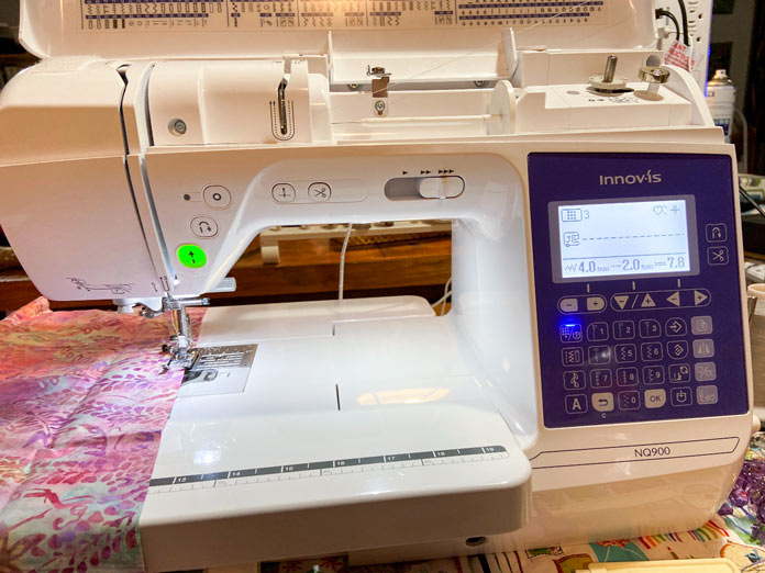 A strip of pink floral batik fabric lays on the extension table of a white and blue Brother NQ900 sewing machine.