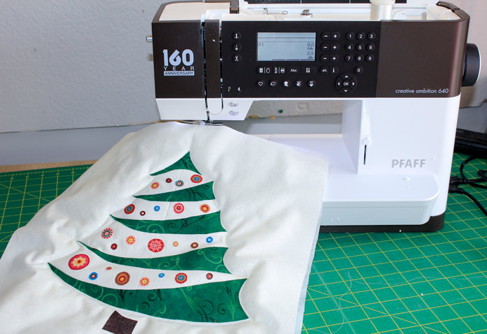 An applique Christmas tree on white fabric under a white and black sewing machine; PFAFF creative ambition 640