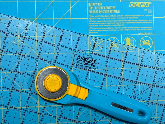 An OLFA 121⁄2″ Square Frosted Acrylic Ruler, Aqua colored OLFA - 12″ x 18″ Double Sided Rotary Mat and an Aqua colored 45mm OLFA Rotary Cutter.