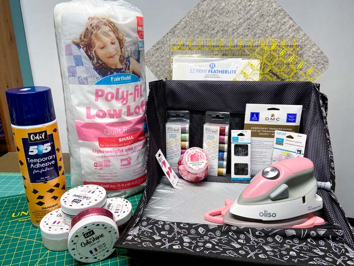 A small pink OLISO iron sits on the folded-out section of a quilting tote along with two packs of perle cotton thread, a jar of buttons, a pack of needles and a package of DMC Embroidery Tracing Paper. A can of Odif 505 Spray, four jars of OdiShine Glitter Gel and a package of Poly-fil Low-Loft Batting sit beside the case.