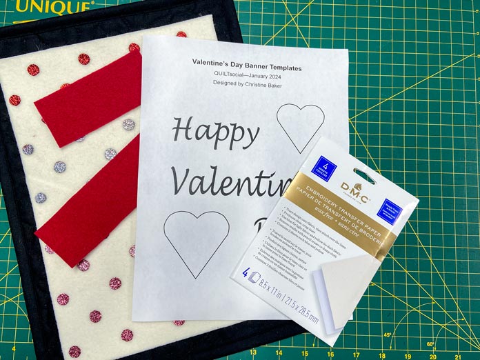 A white, gold, and blue package of embroidery transfer paper is shown sitting on top of a page that says Happy Valentine’s Day as well as some strips of red wool, cream wool with multicolored polka dots and black wool all on a green cutting mat.