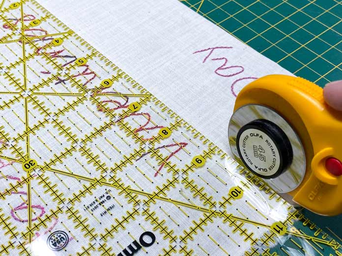 A yellow rotary cutter and ruler cut an embroidered piece of fabric which is sitting on a green cutting mat. CRAFTING ESSENTIALS Bottle of Buttons - Pink Tones - 75g (2.6oz), HEATNBOND EZ Print Feather Lite 10 pcs - 22 x 28cm (81⁄2″ x 11″), SULKY Cotton Petites 6 Spool Thread Set - Rosewood Manor Assortment, UNIQUE Hook & Loop Sew-On Tape - 19mm x 10m (3⁄4″ x 11yd) - Black, OLFA RTY-2/DX - Deluxe Ergonomic Handle Rotary Cutter 45mm, OMNIGRID Ruler - 6″ x 12″, ELAN Novelty 2-Hole Button - Rose - 18mm (3⁄4″) - Heart - 3 count, UNIQUE SEWING Chenille Needles - sizes 18/24 - 6pcs, ODIF OdiShine Glitter Gel, DMC Embroidery Tracing/Transfer Paper, UNIQUE QUILTING Clever Clips Small - 12 pcs, free tutorial.
