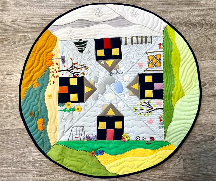A circular table topper with 4 seasons depicted, 4 houses and 4 landscapes with seasonal trees, flowers, snow leaves, pumpkins, a quilt on a clothesline, sports balls and a swing set.