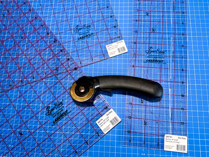 A black rotary cutter on top of three Sew Easy Rulers measuring 15½” x 15½”, 12½” x 12½” and 4½” x 14”; Hemline Gold 45mm Rotary Cutter