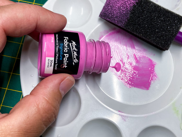 A hand holding a small pink and black bottle pours paint in the center of a white plastic painter’s palette; Mont Marte Round Plastic Palette 6¾”, Mont Marte Signature Fabric Paints