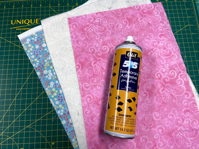 An orange and blue can of Odif 505 Temporary Adhesive Fabric Spray sits on top of two pieces of fabric and a piece of batting. The top fabric has a pink butterfly print and the bottom fabric is a blue floral. There is a green cutting board in the background.