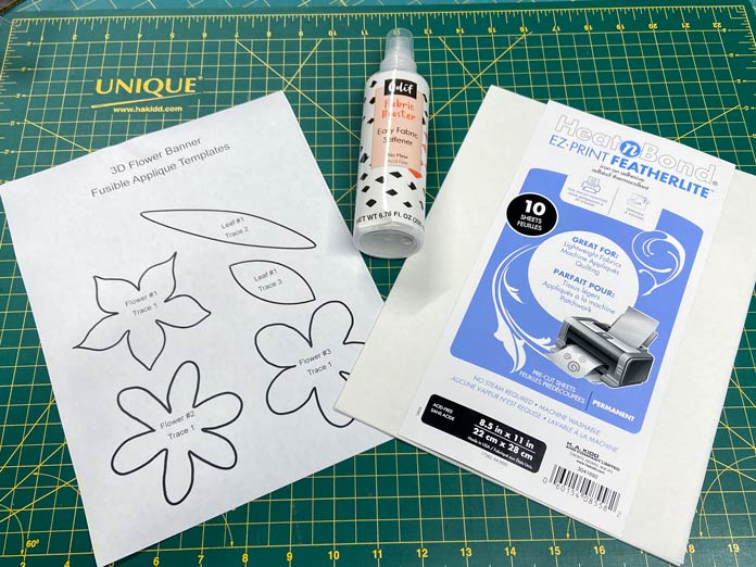 A white paper with outlines of flowers and leaves sits on a green cutting mat beside a purple and white package of HeatnBond EZ Print Featherlite and a spray bottle of Odif Fabric Booster.