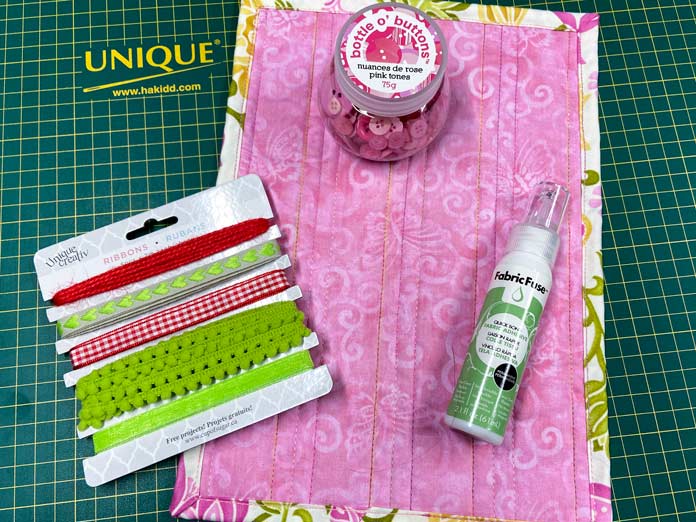 A package of red and green ribbons, a small jar of pink buttons and a gray and white bottle of Fabric Fuse by HeatnBond sit on top of a small pink quilt, all on a green cutting mat; Unique Creativ Trim Pack – Red and Green