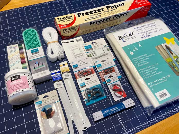 A package of webbing, a box of freezer paper, tote bag hardware, a package of foam stabilizer and other sewing notions sit on top of a blue cutting mat.