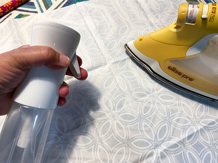 A hand is shown holding a white spray bottle over top of a white fabric that is being pressed with a yellow iron; Odif OdiCoat Gel Coating – 250ml, BEST PRESS Starch Alternative - 499mL (16.9 oz.) - Scent Free, BEST PRESS Spray and Misting Bottle - 295mL (10 fl. oz.) – Empty, Oliso Pro TG1600 Pro Plus Smart Iron, UNIQUE QUILTING Double Sided Applique Pressing Sheet - Brown - 46 x 46cm (18″ x 18″), Bosal In-R-Form Single Sided Fusible Foam Stabilizer - 45.7 x 147.3 cm (18″ x 58″), OLFA SCS-1 - 5″ Stainless Steel Serrated Edge Scissors, free quilting tutorials