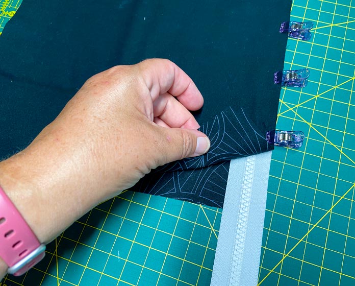 A hand is shown folding back the corner of a black fabric rectangle to show a zipper and another black fabric underneath. Purple clips hold the layers together. There is a green cutting board in the background.