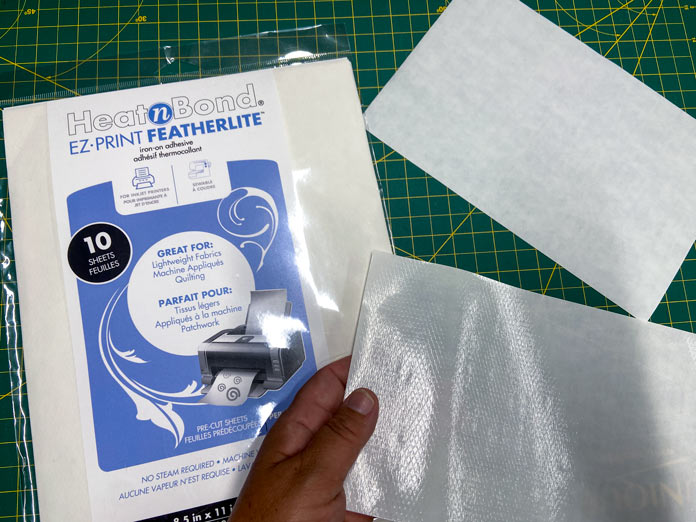 A hand holds a sheet of HeatnBond EZ PRINT FEATHERLITE so that the adhesive side is visible. A Package of HeatnBond EZ PRINT FEATHERLITE and another sheet of the fusible web with the paper side up can be seen in the background.