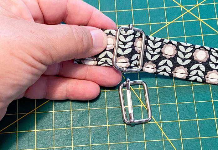 A hand holds a multicolored strap that has been threaded through a silver buckle. A second buckle sits on top of a green cutting mat that is in the background.