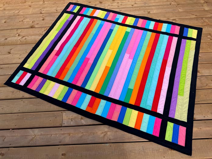 A strip quilt measuring 57” x 70” made with strips of black and bright colors. Hemline Gold Multi-Use Craft Bag (28.5cm x 36.5cm x 17.8cm), Hemline Gold Sewing Kit, Hemline Gold Quilters 4-in-1 Multi-Mat (30cm x 24cm), Hemline Gold Quilters Clips (Pack of 30), Hemlione Gold Retractable Tape Measure - 150cm/60, oliso M3Pro Project Iron – Coral, OLFA 45mm RTY-2/DX/MAG Ergonomic Rotary Cutter – Magenta, OLFA RM-MG - 24″ x 36″ Double Sided Rotary Mat, UNIQUE Sewing Tools Holder, OLFA QR-6x12 - 6″ x 12″ Frosted Acrylic Ruler, OLFA QR-6x24 - 6″ x 24″ Frosted Acrylic Ruler, Fairfield Quilter's 80/20 Quilt Batting - 229 x 274cm (90″ x 108″)