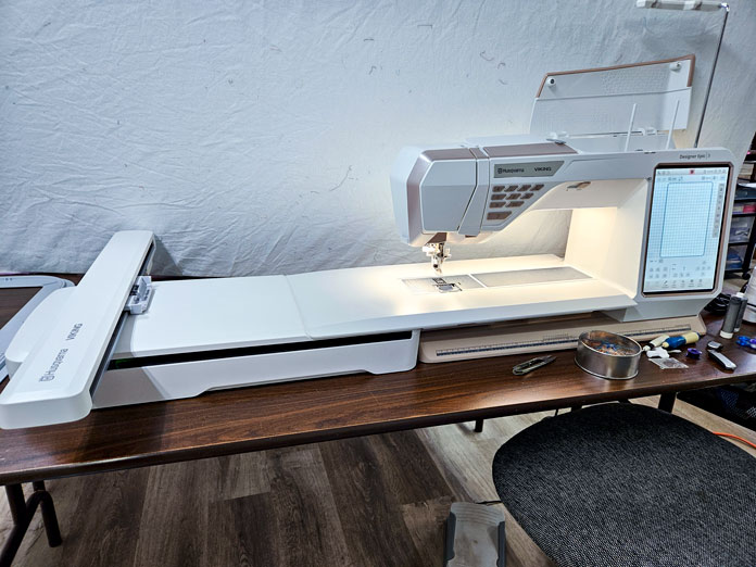 A white and gold embroidery machine with an embroidery unit on a long wooden table; Husqvarna VIKING DESIGNER EPIC 3 sewing and embroidery machine, Husqvarna VIKING 6in/15.2cm Applique Scissor, Husqvarna VIKING Open Toe Presser Foot for IDF System, Husqvarna VIKING Mega Turnable Hoop 450 mm x 450 mm, Husqvarna VIKING Tear-a-Way Extra Wide Stabilizer 24" x 10 Yards, Husqvarna VIKING Cut-a-Way Extra Wide Stabilizer 24” x 10 Yards, SULKY Slimline Metallic Dream Assortment