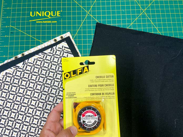 A hand is holding a yellow package that contains an OLFA Chenille Cutter. In the background, some black and white fabrics and a black fabric sit on a green cutting mat. Oliso M2Pro Mini Project Iron, FAIRFIELD Low-Loft Quilt Batting - Crib - 114 x 152cm (45″ x 60″), UNIQUE QUILTING Clever Clips, OLFA CHN-1 - Chenille / Textile Cutter, UNIQUE CREATIV Trim Pack - Black & White, FABRIC CREATIONS Burlap Fabric - Oyster - 45 x 61cm (18″ x 24″), FABRIC CREATIONS Faux Fur Fabric - Zebra - 45 x 76cm (18″ x 30″), UNIQUE SEWING Double Adjust Parachute Buckle - Plastic - 25mm (1″) White, CRAFTING ESSENTIALS Bottle of Buttons - Pink Tones - 75g (2.6oz), UNIQUE SEWING Invisible Magnetic Snaps - Sew-On - 12mm (1⁄2″) - 2 sets, UNIQUE QUILTING Stitch-N-Steam - 157 x 45cm (62″ x 18″), FABRIC CREATIONS Fabric Bundle (5pcs) - Metallic Gold - 45 x 53cm (18in x 21in), Oliso Pro TG1600 Pro Plus Smart Iron, UNIQUE QUILTING Clever Clips Large - 12pcs, UNIQUE SEWING 2-in-1 Dual-Tip Wash-out/Air Erasable Marking Pen - Fine, OMNIGRID Ruler - 121⁄2″ x 121⁄2″ (31.75 x 31.75cm), sewing tutorial