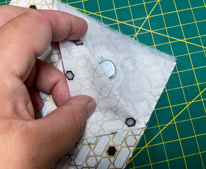 A hand holds a small quilt sandwich made of white and gold fabric. A small magnet sits on the batting, and a green cutting mat can be seen in the background. UNIQUE SEWING Self-Gripping Fastener Strip - 25cm (10″) - White, UNIQUE Webbing 25mm x 10m (1″ x 11yd) - White, ELAN Trench Buckle - 45mm (13⁄4″) - Brown, UNIQUE CREATIV Trim Pack - Black & White, UNIQUE SEWING Invisible Magnetic Snaps - Sew-On - 12mm (1⁄2″) - 2 sets,  SOFTKUT Pinking Shears (Scissors) - 81⁄2″ (21.6cm), COSTUMAKERS General Purpose Closed End Zipper 20cm (8″) - White - 1700, UNIQUE CREATIV Trim Pack - Red & Green, OLFA RTY-2/DX - Deluxe Ergonomic Handle Rotary Cutter 45mm, UNIQUE QUILTING Clever Clips Small - 12 pcs, UNIQUE SEWING Double Adjust Parachute Buckle - Plastic - 25mm (1″) - White, CRAFTING ESSENTIALS Bottle of Buttons - Pink Tones - 75g (2.6oz), UNIQUE Braided Elastic 3mm x 3.7m - White, free tutorial