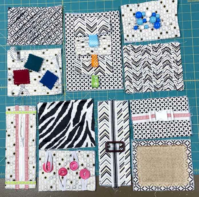 11 different textured and activity quilt blocks in black, white, and gold fabrics with colorful accents sit on a green cutting mat. UNIQUE SEWING Invisible Magnetic Snaps - Sew-On - 12mm (1⁄2″) - 2 sets, COSTUMAKERS General Purpose Closed End Zipper 20cm (8″) - White - 1700, UNIQUE SEWING Double Adjust Parachute Buckle - Plastic - 25mm (1″) – White, UNIQUE QUILTING Clever Clips Small - 12 pcs, CLOVER 7812 Roll & Press, UNIQUE QUILTING Wool Pressing Mat - 18″ x 24″ - Grey, OLISO M2Pro Mini Project Iron with Solemate, CLOVER 240 - Fork Pins - .56mm, CLOVER 464/18 - Bias Tape Maker - 18mm (3⁄4″), Odif 505 Temporary Quilt Basting Adhesive Fabric Spray - 156g, FAIRFIELD Low-Loft® Quilt Batting - Crib - 114 x 152cm (45″ x 60″), free tutorial