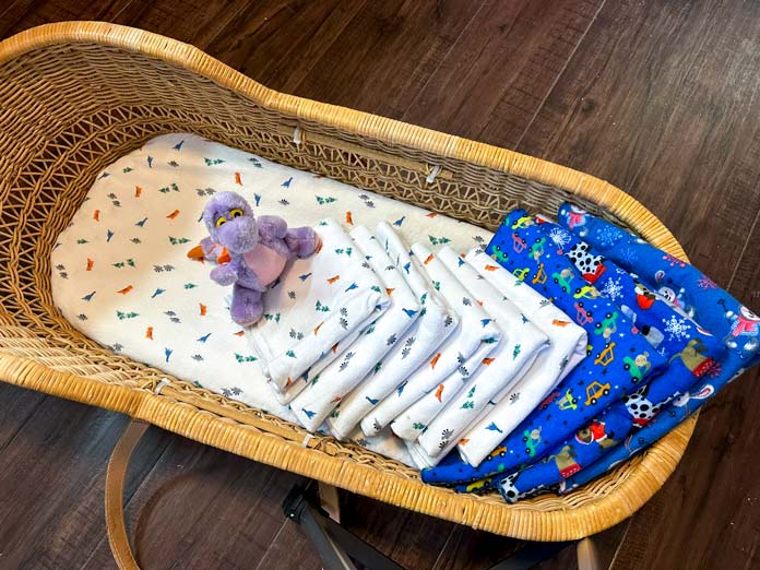 A wicker bassinet made up with white flannel sheets, matching white flannel receiving blankets laid on top, with extra large blue receiving blankets, snuggled in with a small purple dinosaur stuffy