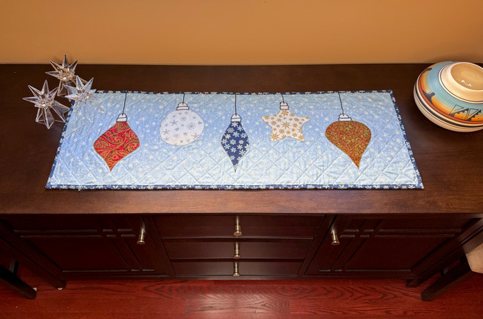 A table runner with 5 Christmas decorations appliqued on a blue snowflake fabric. The table runner is on a dining room sideboard flanked by three candle holders on one side and a pottery bowl on the other. Gütermann Dekor Metallic Thread 200m – Gold, Gütermann Dekor Metallic Thread 200m - Col. 41, Gütermann Dekor Metallic Thread 200m – Red, Gütermann Dekor Metallic Thread 200m - Christmas Green, Gütermann Dekor Metallic Thread 200m - Col. 1005, Gütermann Denim Thread 100m – Black, UNIQUE Baby Rick Rack 3mm x 3.7m – Scarlet, UNIQUE Medium Rick Rack 6mm x 2.3m - Emerald Green, UNIQUE Jumbo Rick Rack 10mm x 2.3m - Metallic Gold, SCHMETZ #1743 Metallic Needles Carded - 80/12 - 5 count, SCHMETZ #1752 Metallic Needles Carded - 90/14 - 5 count, LDH 8½“ Craft Scissors – Purple, HeatnBond Lite Iron-On Adhesive - 43cm x 30m (17″ x 32 ¾yds) bolt, HeatnBond Stitch n Sew Tear-Away Stabilizer - 50.8cm x 22.8m (20″ x 25yds), oliso M3Pro Project Iron – Coral, OLFA 45mm RTY-2/DX/MAG Ergonomic Rotary Cutter – Magenta, UNIQUE QUILTING Wool Pressing Mat - 18″ x 24″ - Grey 