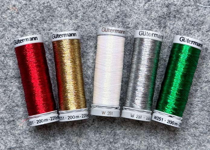 5 spools of Gütermann Metallic threads, one each of red, gold, white, silver and green; Gütermann Dekor Metallic Thread 200m – Gold, Gütermann Dekor Metallic Thread 200m - Col. 41, Gütermann Dekor Metallic Thread 200m – Red, Gütermann Dekor Metallic Thread 200m - Christmas Green, Gütermann Dekor Metallic Thread 200m - Col. 1005, Gütermann Denim Thread 100m – Black, UNIQUE Baby Rick Rack 3mm x 3.7m – Scarlet, UNIQUE Medium Rick Rack 6mm x 2.3m - Emerald Green, UNIQUE Jumbo Rick Rack 10mm x 2.3m - Metallic Gold, SCHMETZ #1743 Metallic Needles Carded - 80/12 - 5 count, SCHMETZ #1752 Metallic Needles Carded - 90/14 - 5 count, LDH 8½“ Craft Scissors – Purple, HeatnBond Lite Iron-On Adhesive - 43cm x 30m (17″ x 32 ¾yds) bolt, HeatnBond Stitch n Sew Tear-Away Stabilizer - 50.8cm x 22.8m (20″ x 25yds), oliso M3Pro Project Iron – Coral, OLFA 45mm RTY-2/DX/MAG Ergonomic Rotary Cutter – Magenta, UNIQUE Quilting Wool Pressing Mat - 18″ x 24″ - Grey