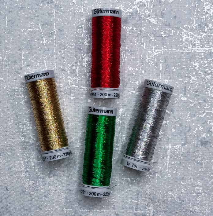 4 spools of Gütermann Metallic Threads, one each of gold, red, green, and silver; Gütermann Dekor Metallic Thread 200m – Gold, Gütermann Dekor Metallic Thread 200m - Col. 41, Gütermann Dekor Metallic Thread 200m – Red, Gütermann Dekor Metallic Thread 200m - Christmas Green, Gütermann Dekor Metallic Thread 200m - Col. 1005, Gütermann Denim Thread 100m – Black, UNIQUE Baby Rick Rack 3mm x 3.7m – Scarlet, UNIQUE Medium Rick Rack 6mm x 2.3m - Emerald Green, UNIQUE Jumbo Rick Rack 10mm x 2.3m - Metallic Gold, SCHMETZ #1743 Metallic Needles Carded - 80/12 - 5 count, SCHMETZ #1752 Metallic Needles Carded - 90/14 - 5 count, LDH 8½“ Craft Scissors – Purple, HeatnBond Lite Iron-On Adhesive - 43cm x 30m (17″ x 32 ¾yds) bolt, HeatnBond Stitch n Sew Tear-Away Stabilizer - 50.8cm x 22.8m (20″ x 25yds), oliso M3Pro Project Iron – Coral, OLFA 45mm RTY-2/DX/MAG Ergonomic Rotary Cutter – Magenta, UNIQUE Quilting Wool Pressing Mat - 18″ x 24″ - Grey