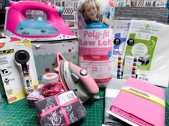 A large iron, mini-iron, package of quilt batting, fabrics, rotary cutter, and other sewing notions sit on top of a green cutting mat