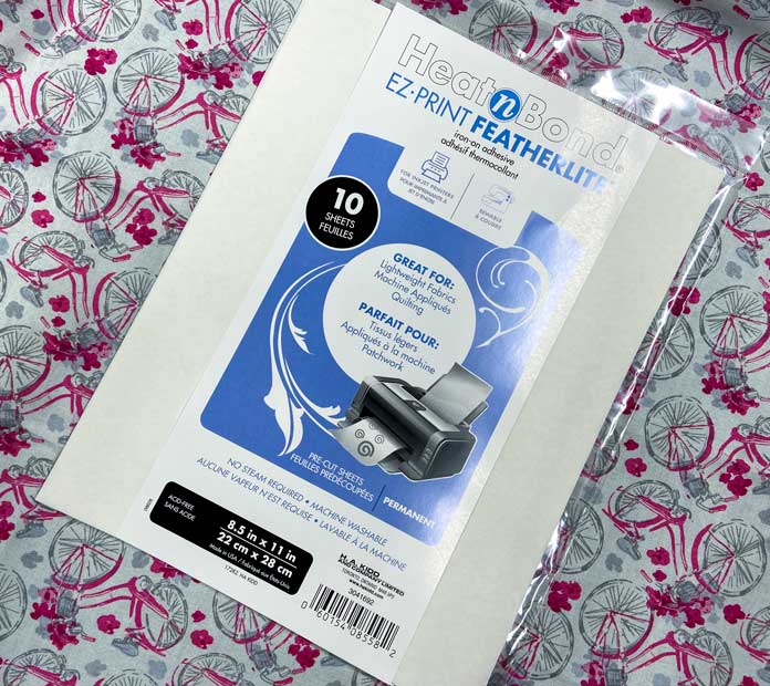 A white and blue package of HeatnBond EZ Print Featherlite Sheets sits on top of some gray and pink fabric that has a bicycle design.