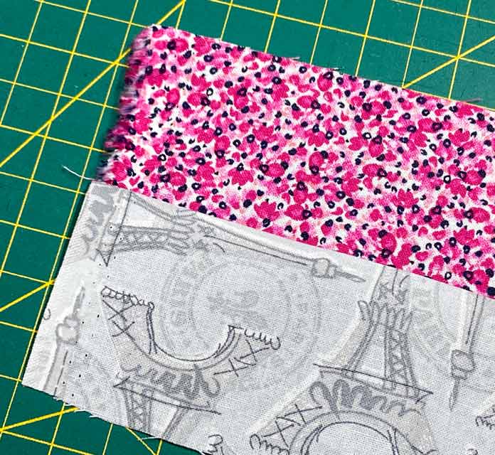 A gray fabric with images of the Eiffel Tower is sewn to a strip of pink mottled fabric. The selvedge ends of the strips are both at the same end of the strip set. A green cutting mat is in the background.