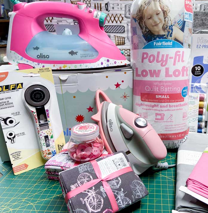 A large iron, mini-iron, package of quilt batting, fabrics, rotary cutter, and other sewing notions sit on top of a green cutting mat.