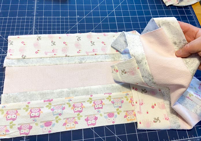 Five strips of pink, white and gray fabrics that have been sewn together and have uneven ends sit on top of a blue cutting mat. Hand holds up the right end to show that the ends of the strips are uneven.