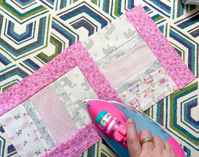 A hand uses a bright pink iron to press the seam between two pink, gray and white quilt blocks. The blocks sit on top of a multicolored ironing board cover.