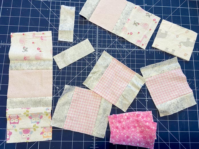 An assortment of 4½" wide block pieces in pink, gray and white fabrics sit on top of a blue cutting mat. 