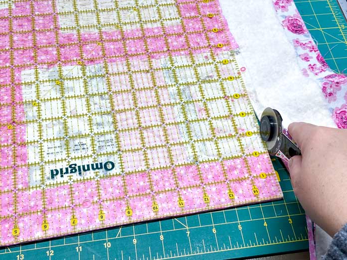 A hand uses a white rotary cutter to trim off the corner of a quilt using a large square ruler. A green cutting mat is in the background.