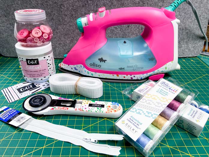 A package of webbing, two packs of threads, an iron, zipper and rotary cutter sit on top of a green cutting mat. 