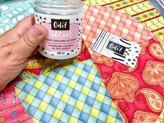 A hand is shown holding a white jar of Odicoat, with two multicolored quilted quilt blocks in the background. A white plastic card that reads 'Odif' lies on top of the quilt blocks.