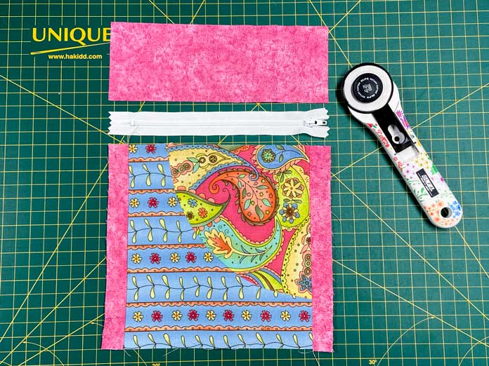 A multicolored quilt block, a zipper, a rectangle of pink fabric, and a white-handled rotary cutter, decorated with a floral design, sit on top of a green cutting mat.