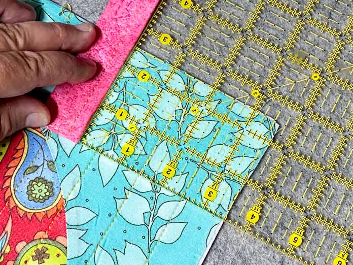 A hand holds a pink strap on top of a multicolored quilted panel. A rotary cutting ruler is on top of the corner of the panel, measuring in 3½" from the side and 2½" from the top. A grey wool pressing mat sits in the background.