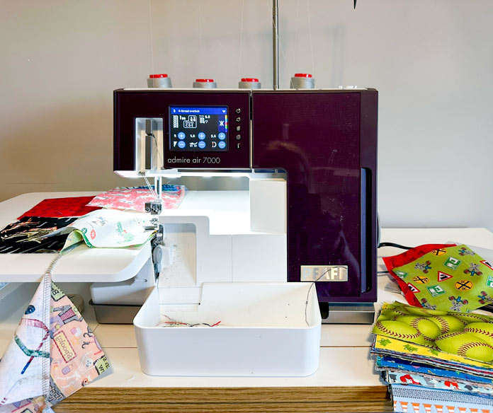 A white and burgundy serger with a white extension table attached. 5” colorful squares being chain pieced into rows, with a pile of squares off to the left.