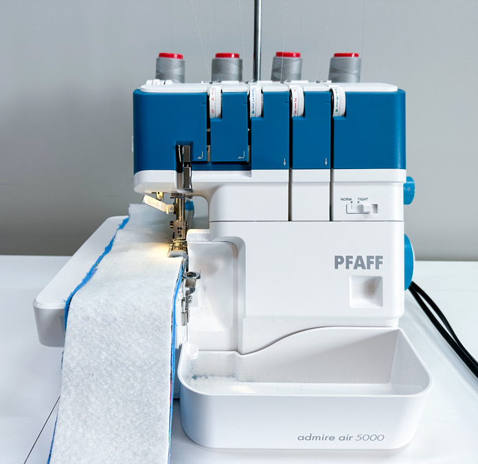 A white and blue serger, set up with 4 spools of grey thread, loaded with a thick sandwich of fabric and batting.