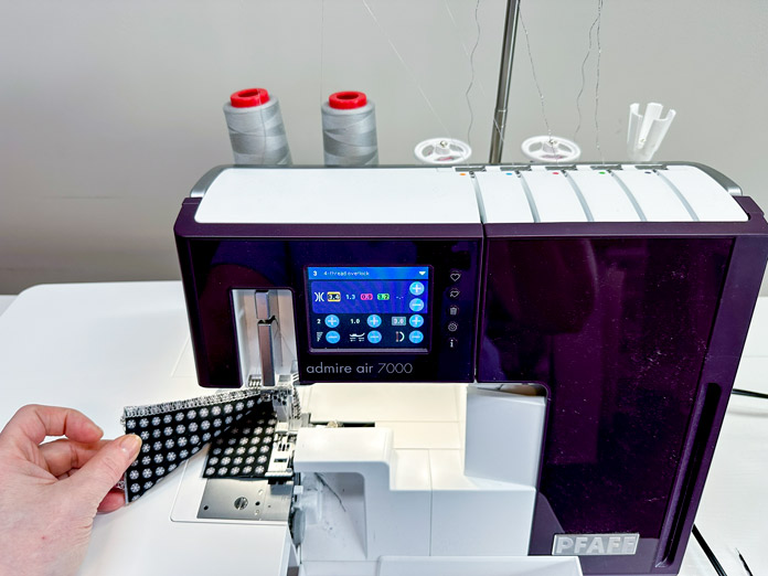 A white and burgundy serger with an extension table showing a bold 4-thread overlock gray stitch on black and silver fabric. The looper threads are thick, gray metallic. 