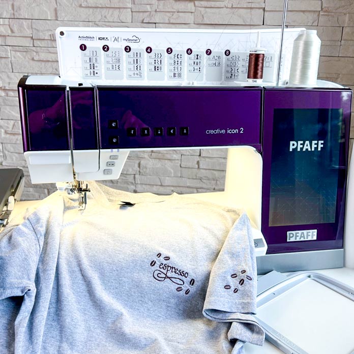 The gray cotton T-shirt features a satin stitch design embroidered on the left chest and sleeve. The design includes the word “espresso” surrounded by scattered coffee beans in brown thread. The machine’s screen and needle arm are visible in the background, with thread spools placed on top. The embroidery hoop is positioned beside the machine.