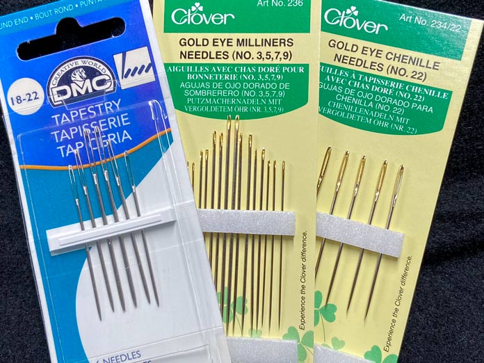 One white and blue package of DMC tapestry needles and two yellow packages of chenille and milliner’s needles on a black background; Clover Gold Eye Milliner Needles (No. 3,5,7,9), DMC Tapestry Needles Size 18-22